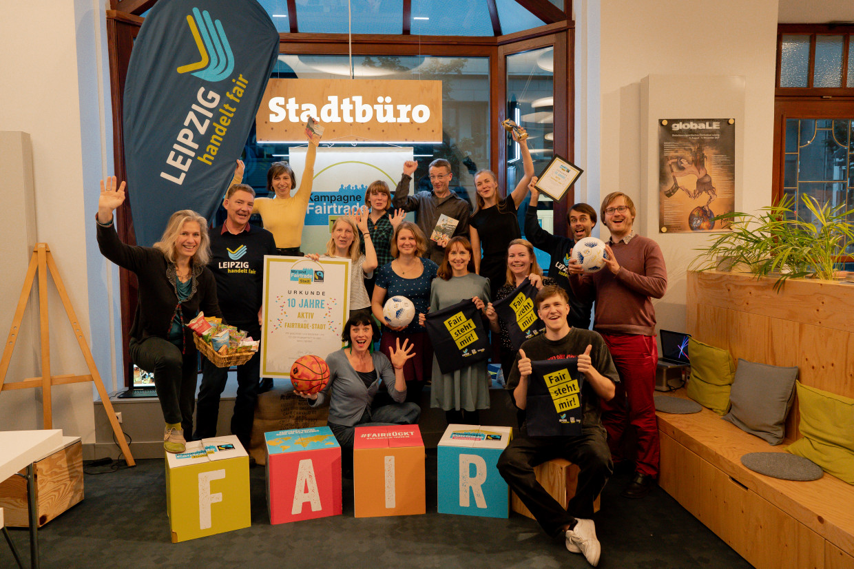 Das Leipzig handelt fair Team mit einem großen Stoffaufsteller, welcher das Logo des Vereins zeigt, stehen vor dem Leipziger Rathaus. Sie schauen freundlich in die Kamera.