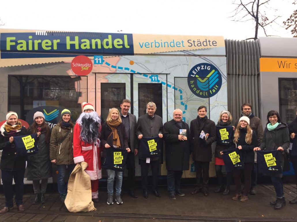 Das Leipzig handelt fair Team mit einem großen Stoffaufsteller, welcher das Logo des Vereins zeigt, stehen vor dem Leipziger Rathaus. Sie schauen freundlich in die Kamera.